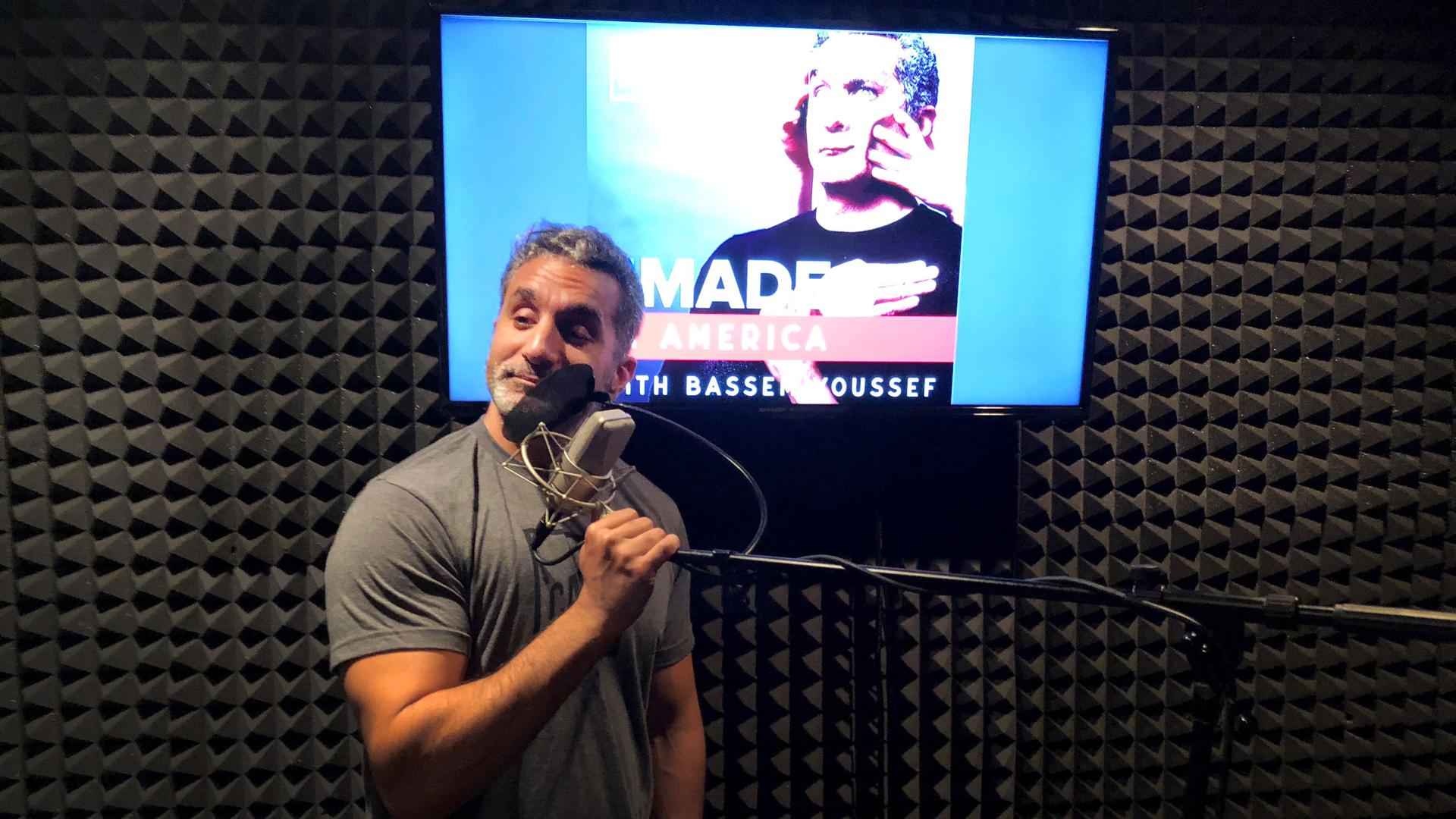 Bassem Youssef stands in front of a microphone in a recording studio. Behind him is a screen lit up with the title of his new podcast: "Remade in America with Bassem Youssef."
