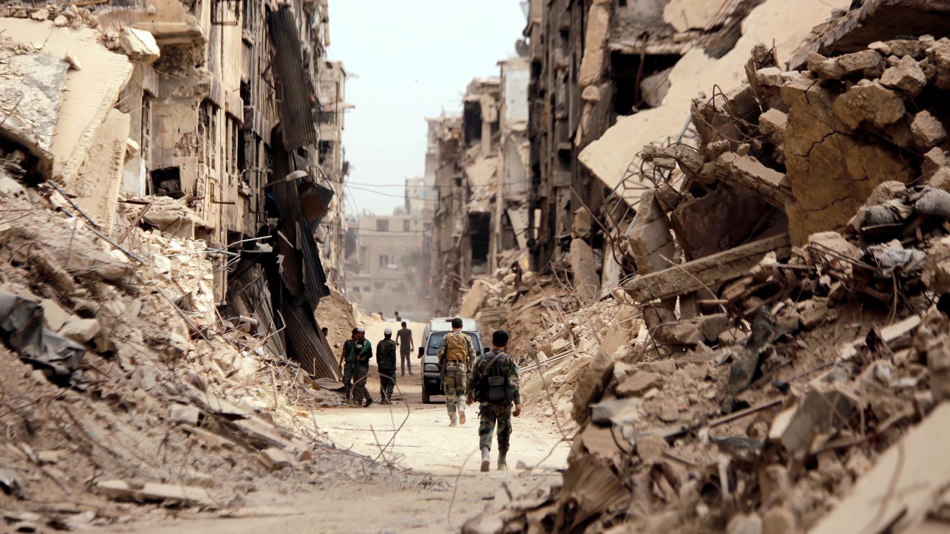 Men in camouflague fatigues walk down a street lined with rubble from destroyed buildings.