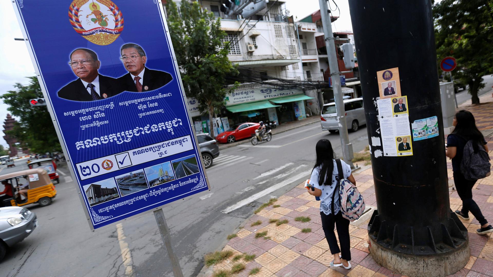 A blue sign showing two politicians stands next to an intersection in a city, where a woman is waiting to cross the street. The signs are written in Cambodian. 