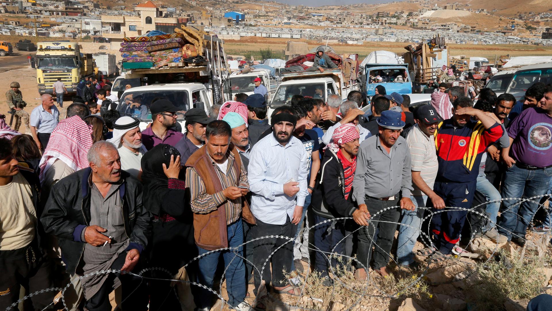 People stand at a line behind a barbed wire fence. Behind them is a line of trucks laden with mattresses and other supplies.