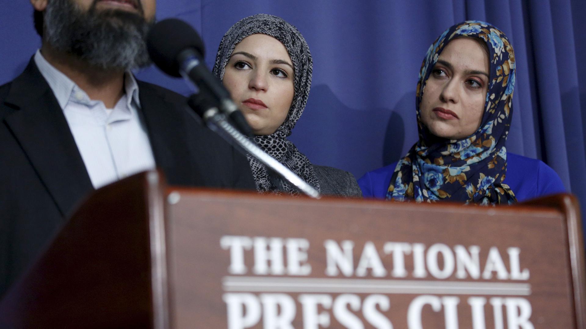 Two women wearing head scarves watch a man at a podium. 