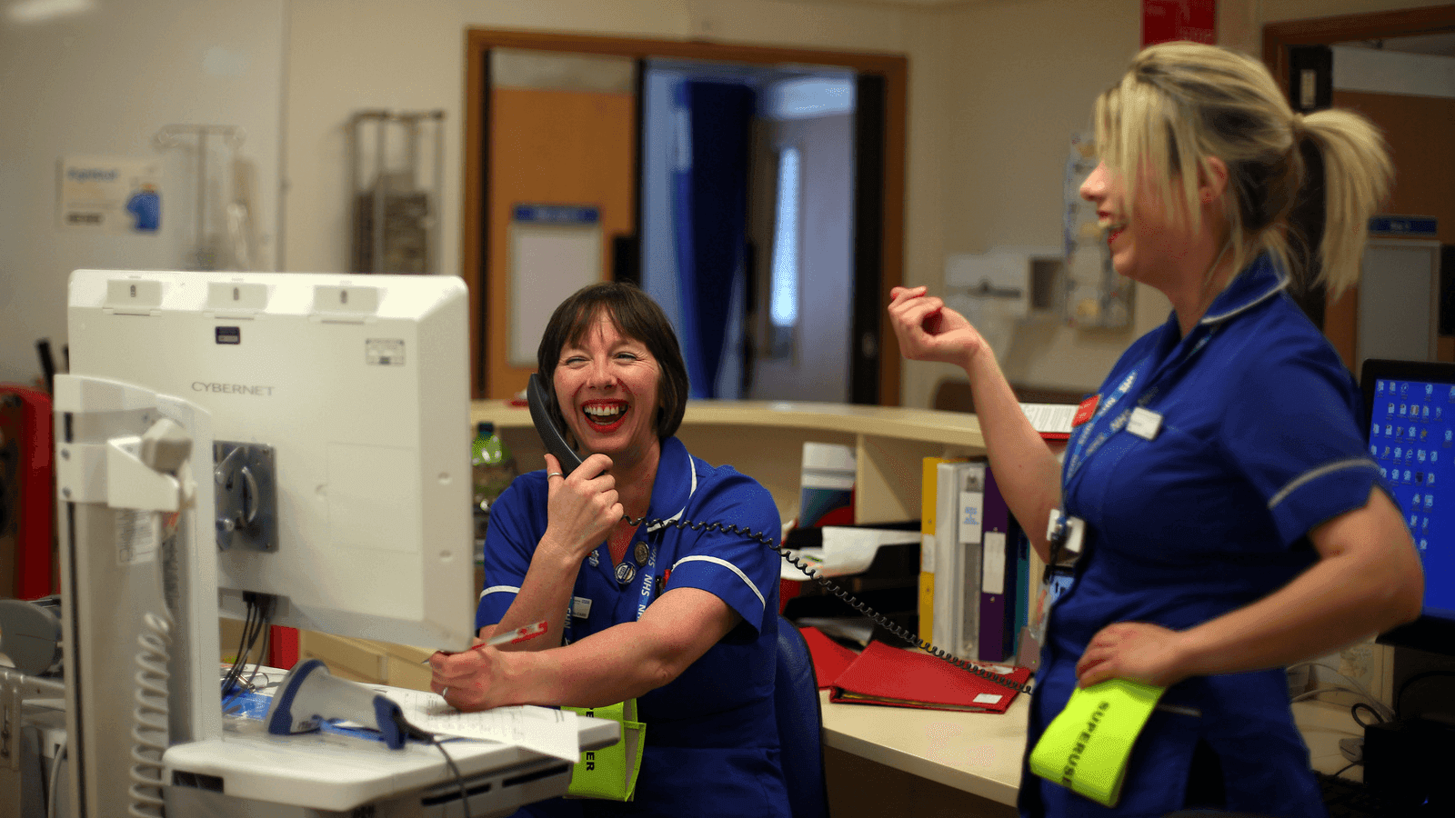 two nurses laugh at a work station in a hospital 