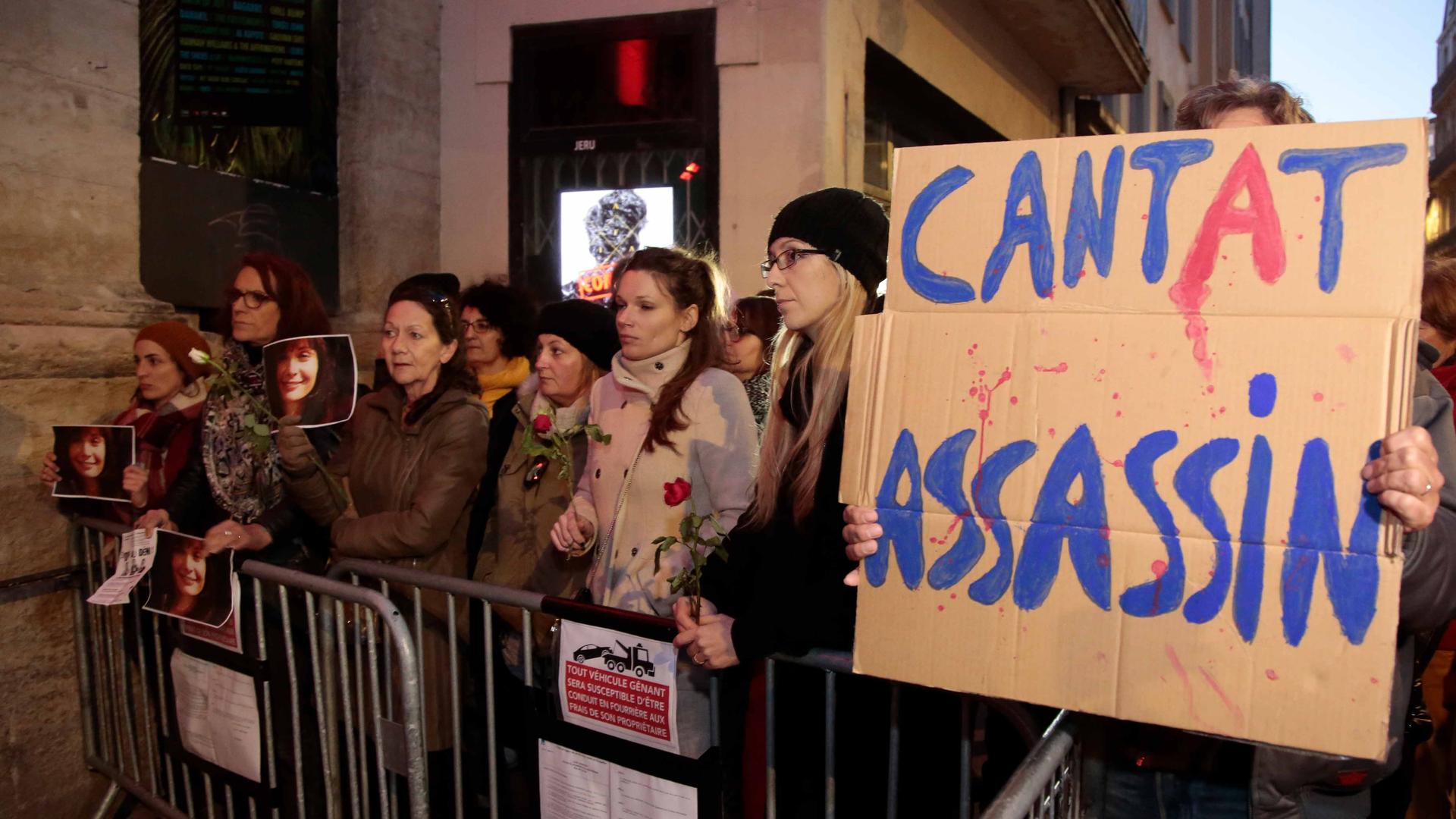 A crowd stands behind a metal gate. One holds a sign that says "Cantat assassin"