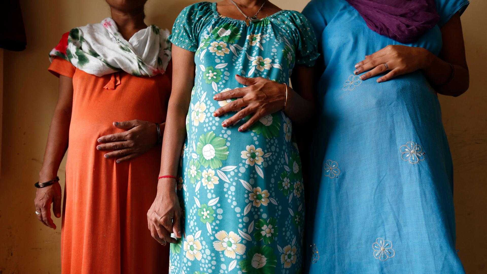 Three women stand in a line with their hands on their visibly pregnant stomachs. Their faces are cropped out of the image. 