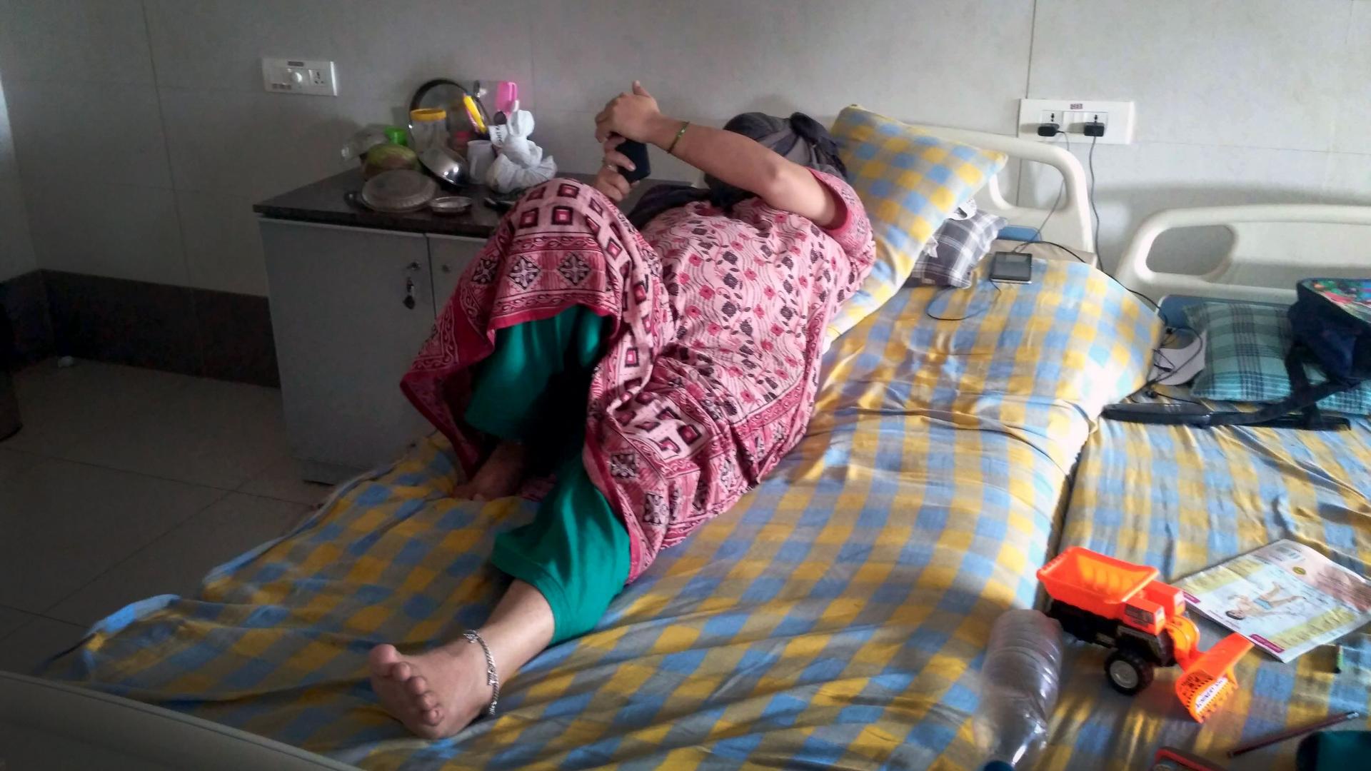 A woman lays on a hospital bed and turns her face away from the camera, reading the cell phone in her hand.
