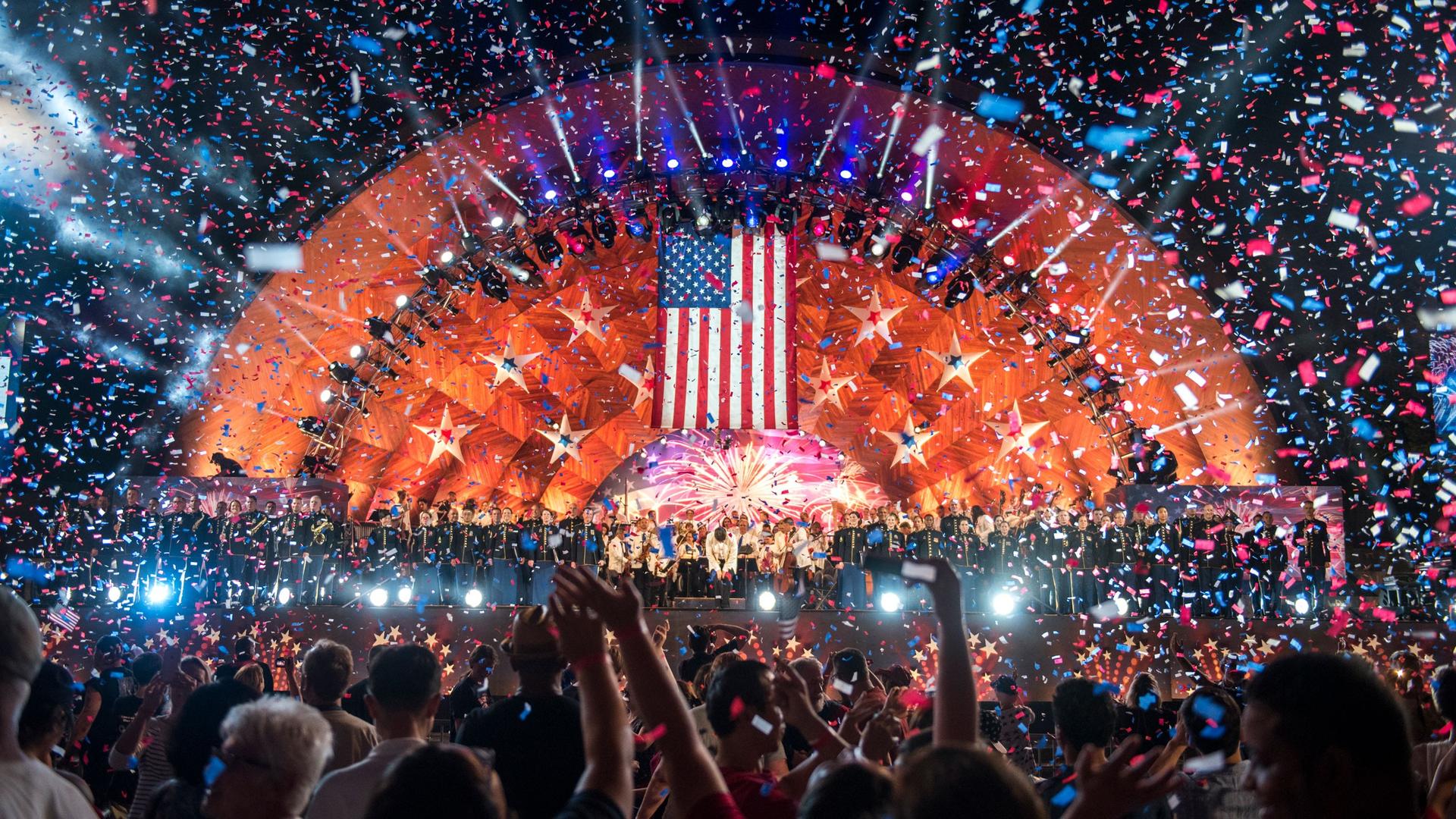 A crowd watches an illuminated bandshell decorated with red and blue stars while confetti falls in the air.