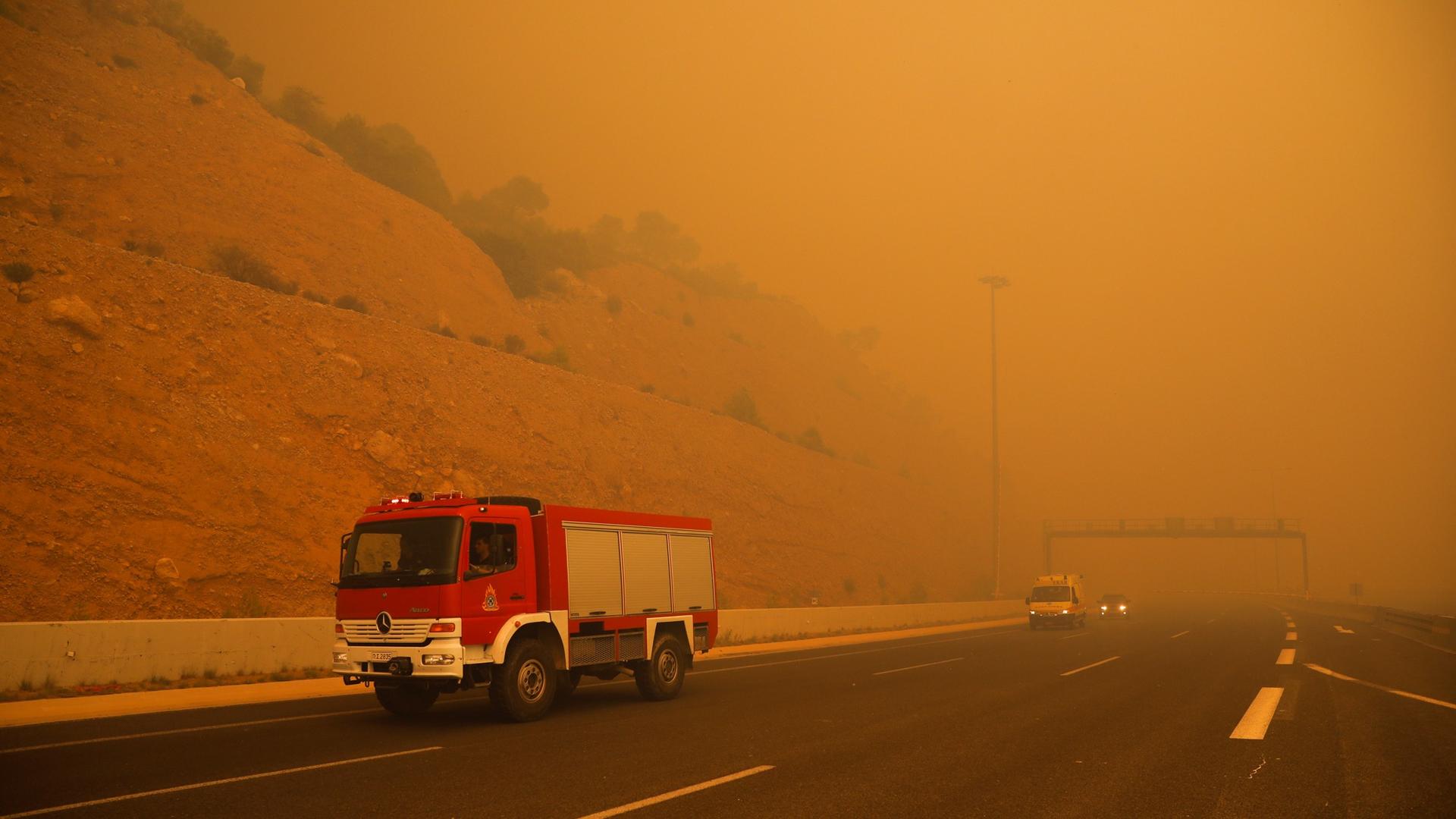 A fire truck drives along a road in Greece amid orange smoke.
