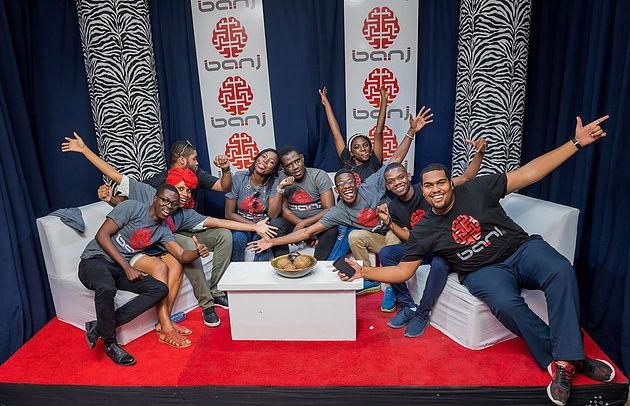 Group sitting on sofa poses for camera with exuberant facial expressions, all wearing Banj T-shirts, red carpet in front, colorful wall behind