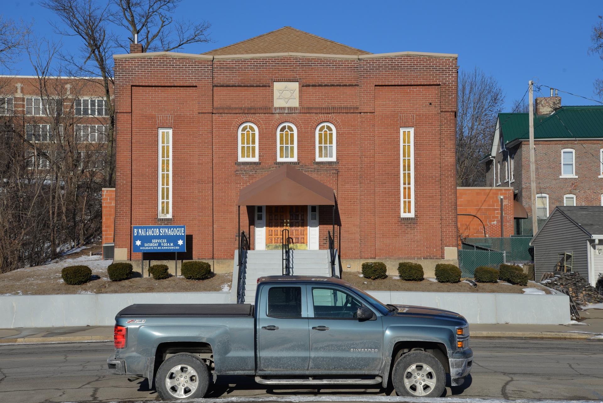 Small brick building with synagogue sign in front, on small street. Truck in foreground.