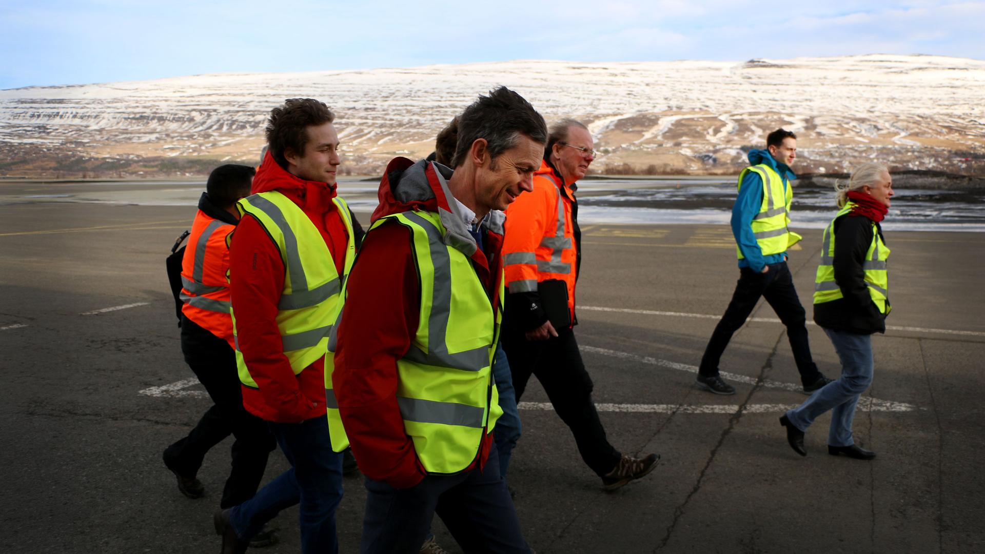 A group of people in bright safety vests walk through the wind on a tarmac.