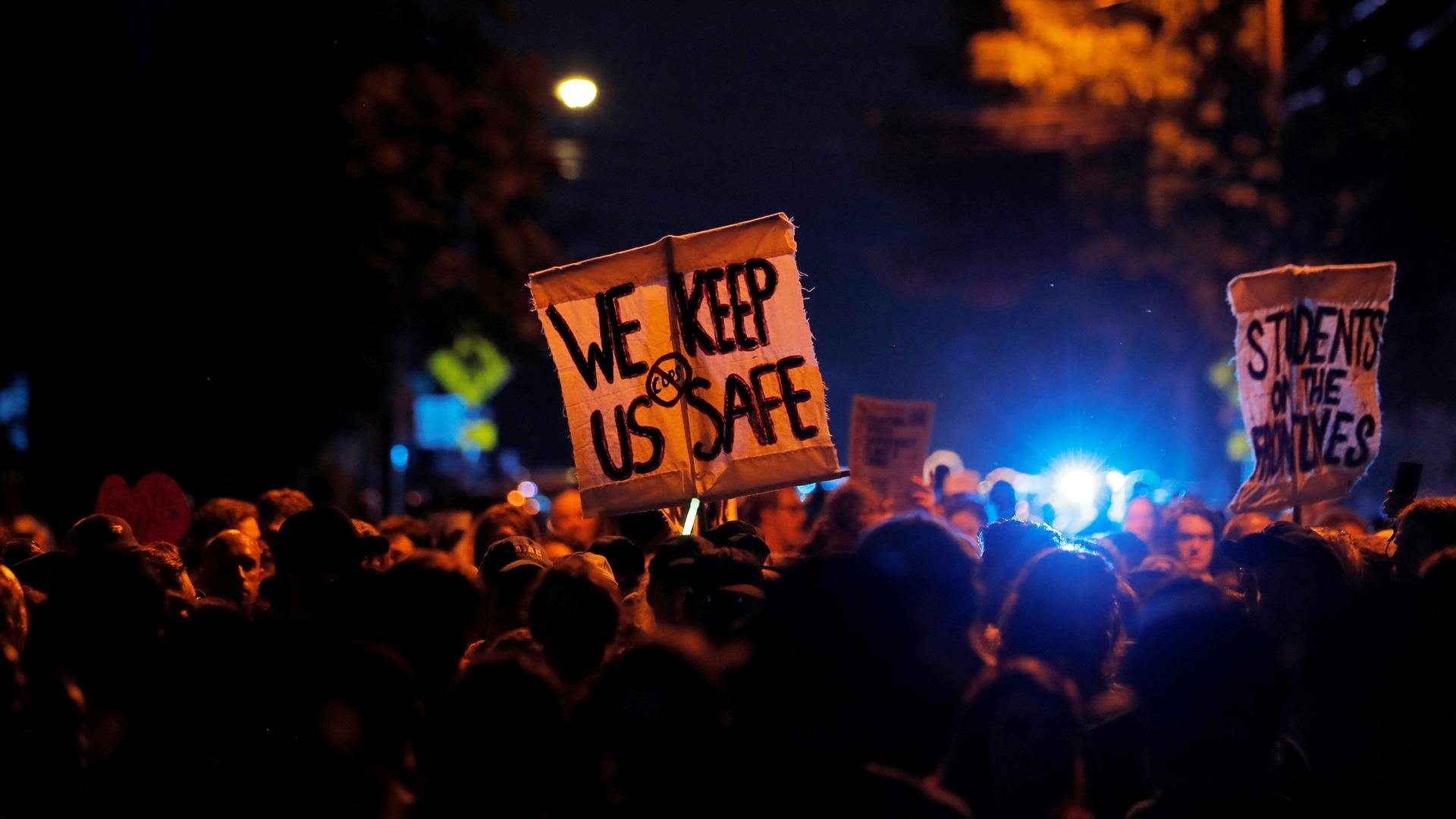A crowd of people is illuminated by the blue of a police light and a sign says 