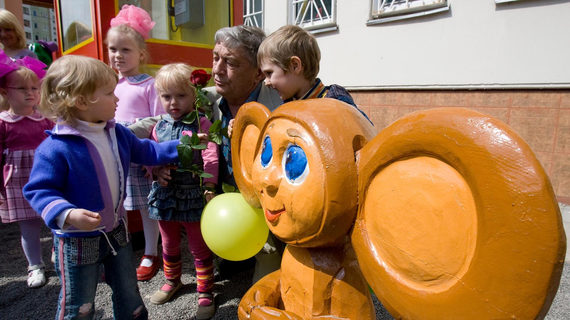 A man poses with small children next to a wooden image of the cartoon character Chebrashka.