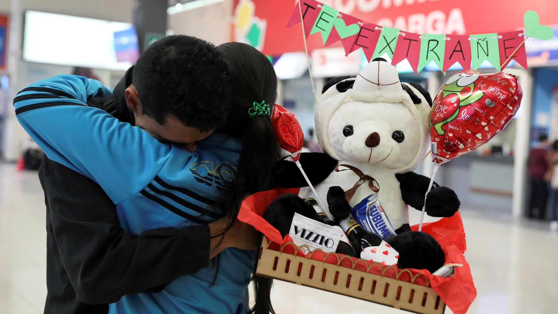 Two people embrace in a bus station. One of them holds a greeting balloon, candy and a stuffed animal.