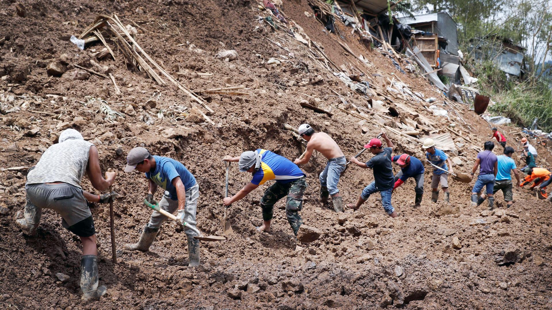 A row of men with shovels stand on a hillside covered in debris and loose mud after a mudslide covered a mining town
