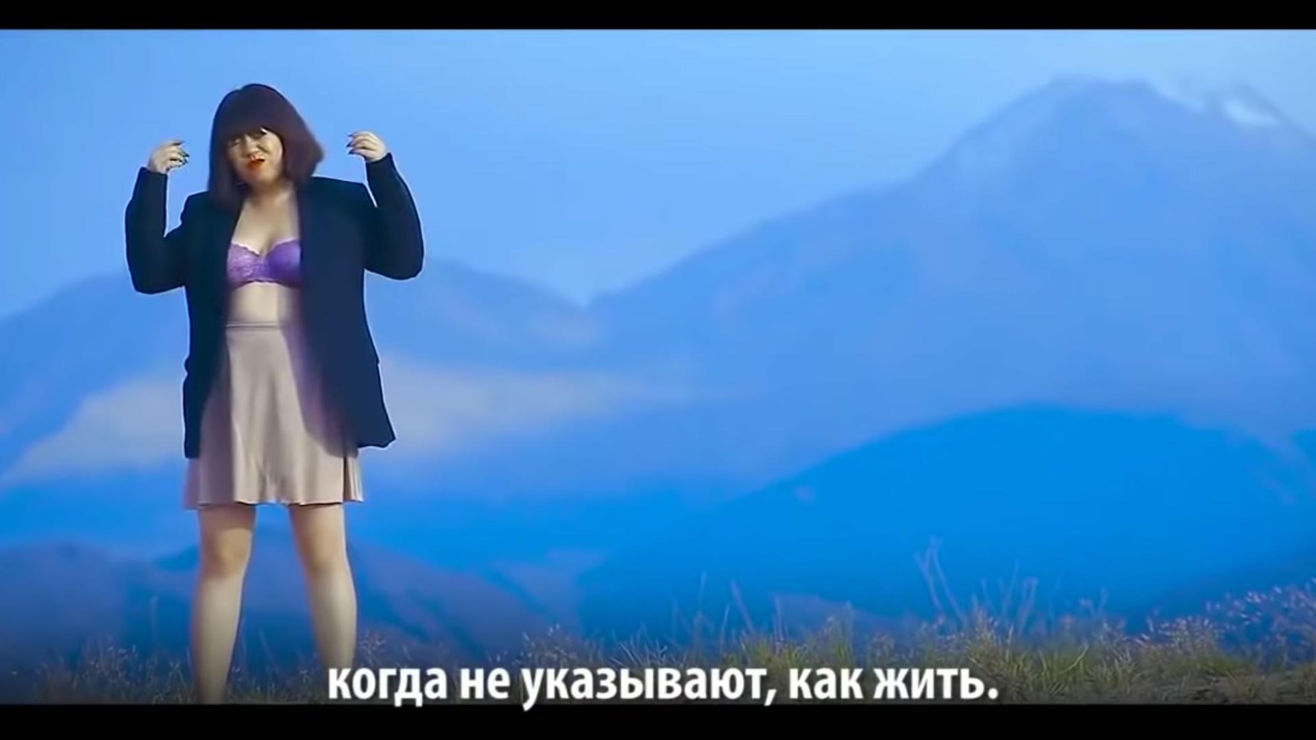 A woman wears a lacy bra and a blazer as she stands in front of a sweeping mountain vista in this screen grab from a music video. Below, a line from the song is spelled out in Kyrgyz. 