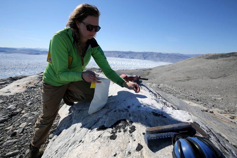 A woman picks something off a rock as a ice field glitters in the background