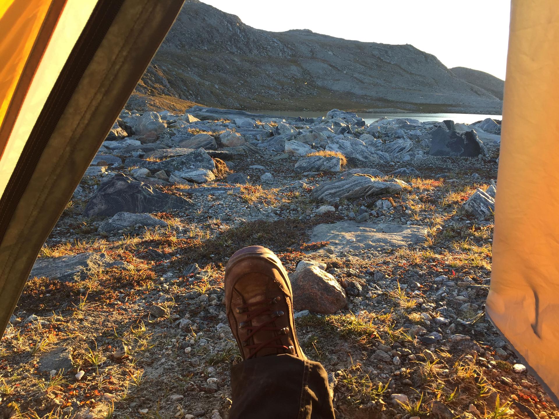 A woman's booted foot is framed by the walls of her tent as the golden sunlight spills over rocky terrain.
