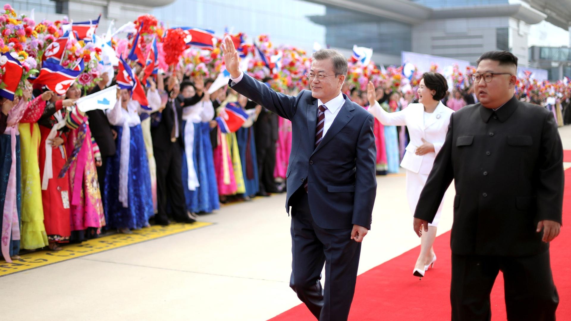 South Korean President Moon Jae-in and North Korean leader Kim Jong-un walk past a welcoming crowd in traditional Korean dress waving flowers and North Korean flags. 