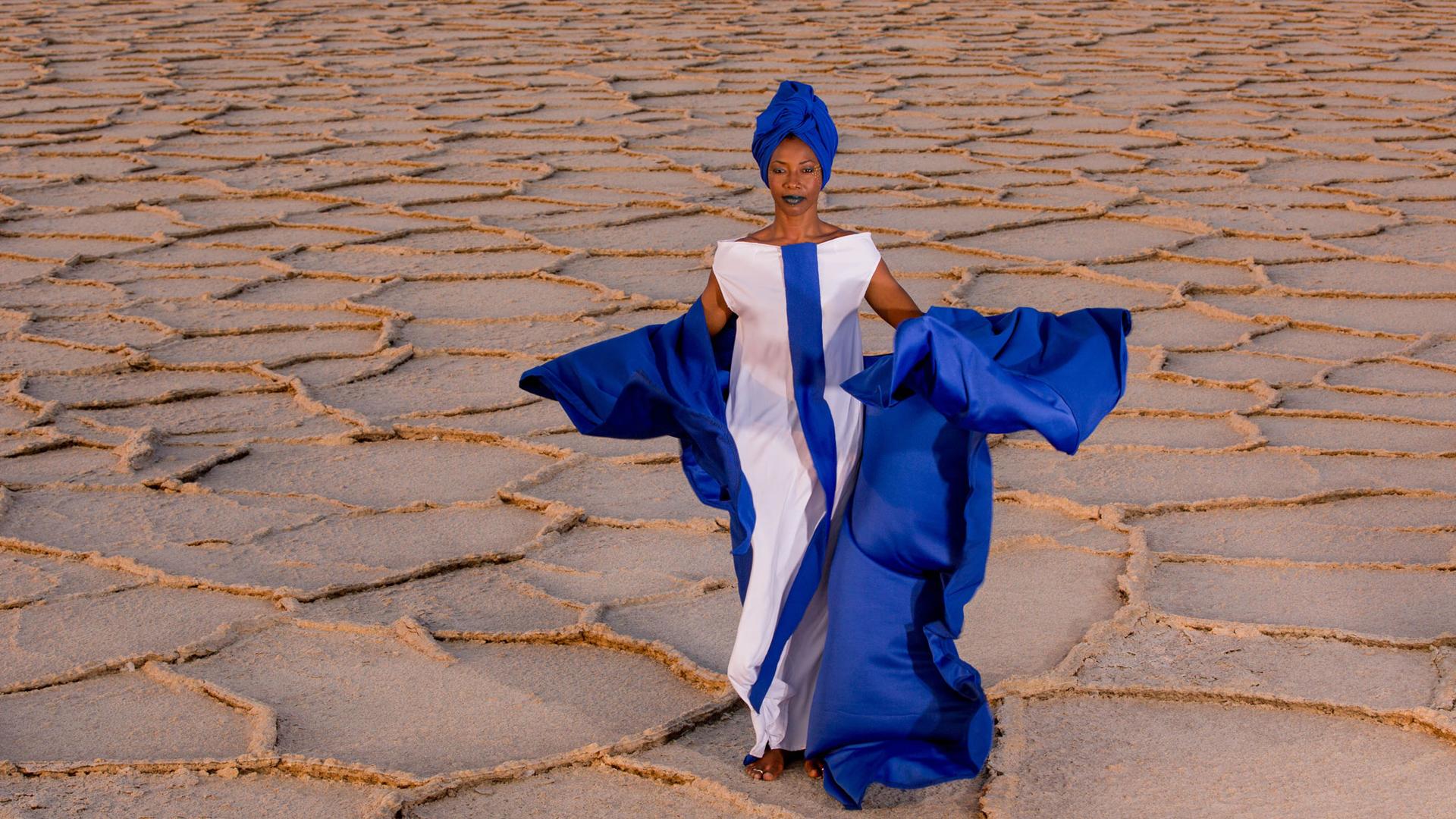 Malian singer Fatoumata Diawara is shown in a bright blue and white dress standing in the desert. 