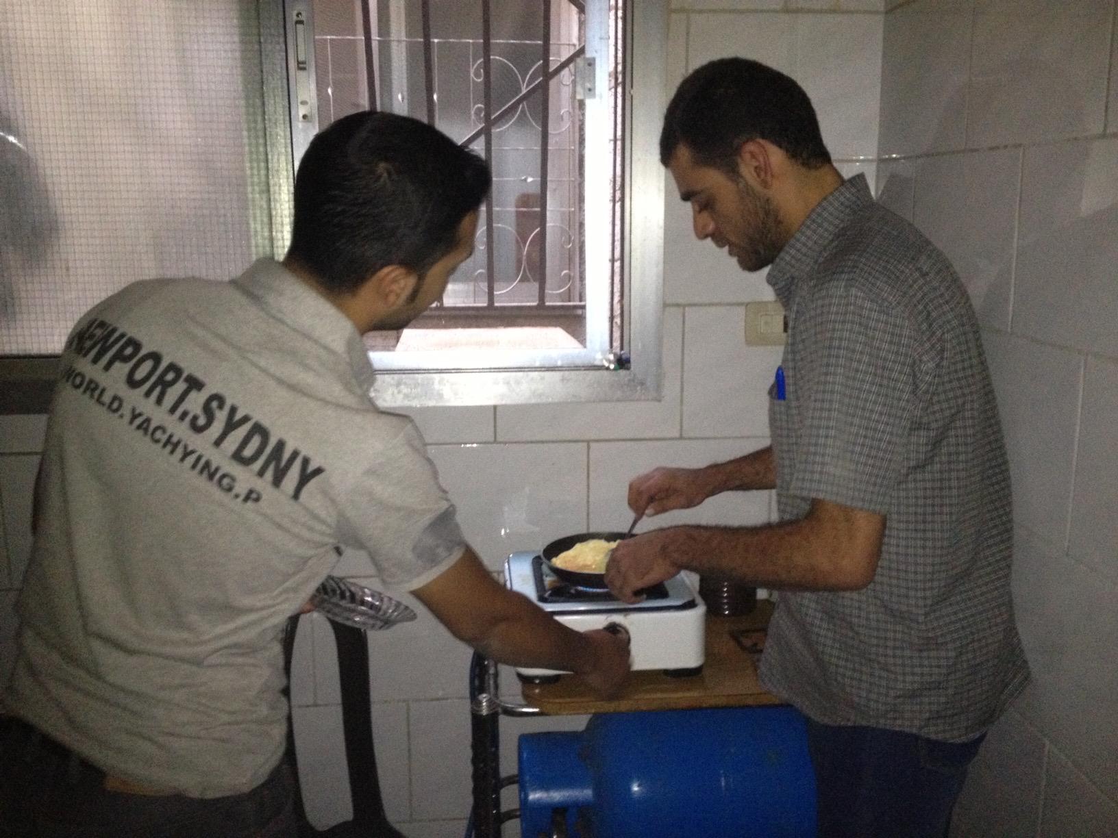 two men standing in a small, dark kitchen making eggs on a hotplate