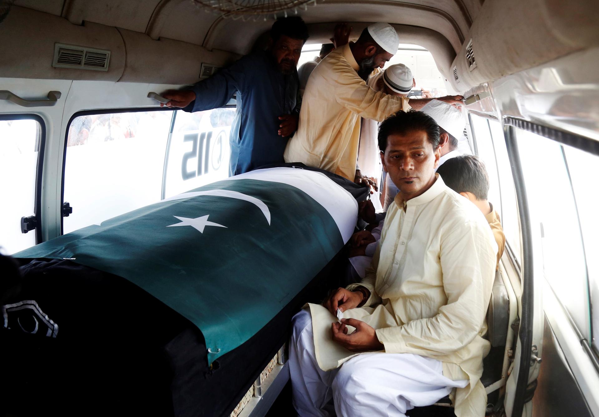 Man inside van sits next to coffin with Pakistan flag draped over, others standing nearby in close quarters
