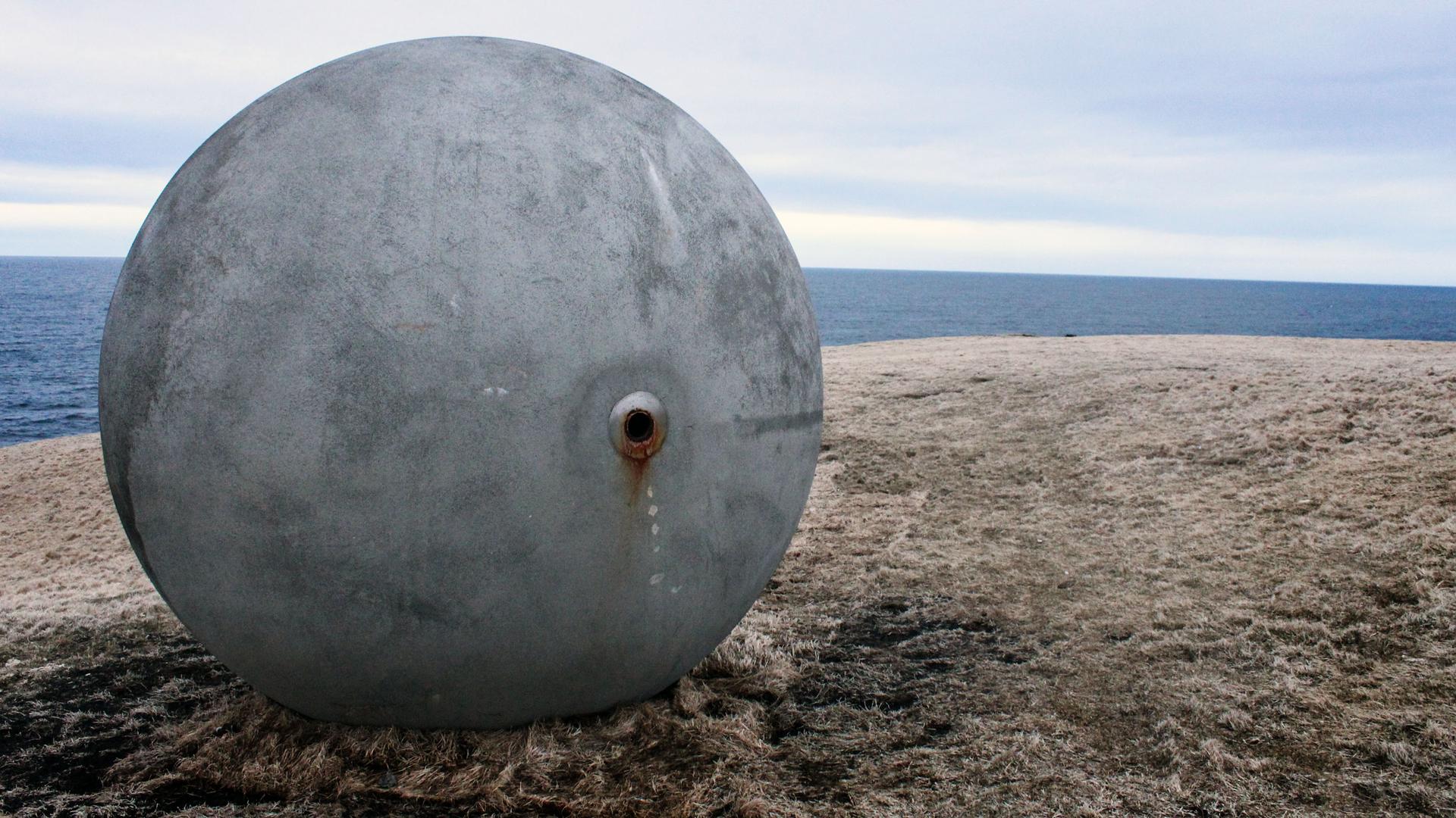 A large grey sphere sits on a grassy hill by a sea