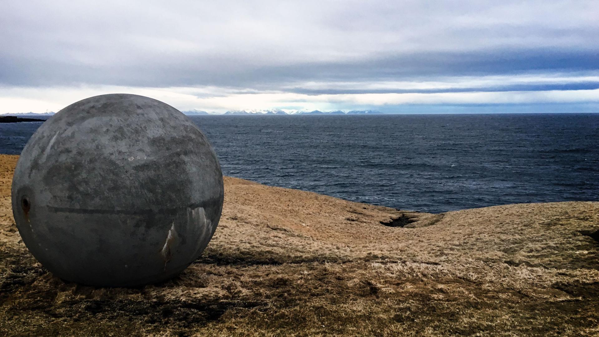 A large concrete sphere sits on a brown hill overlooking a slate blue sea.