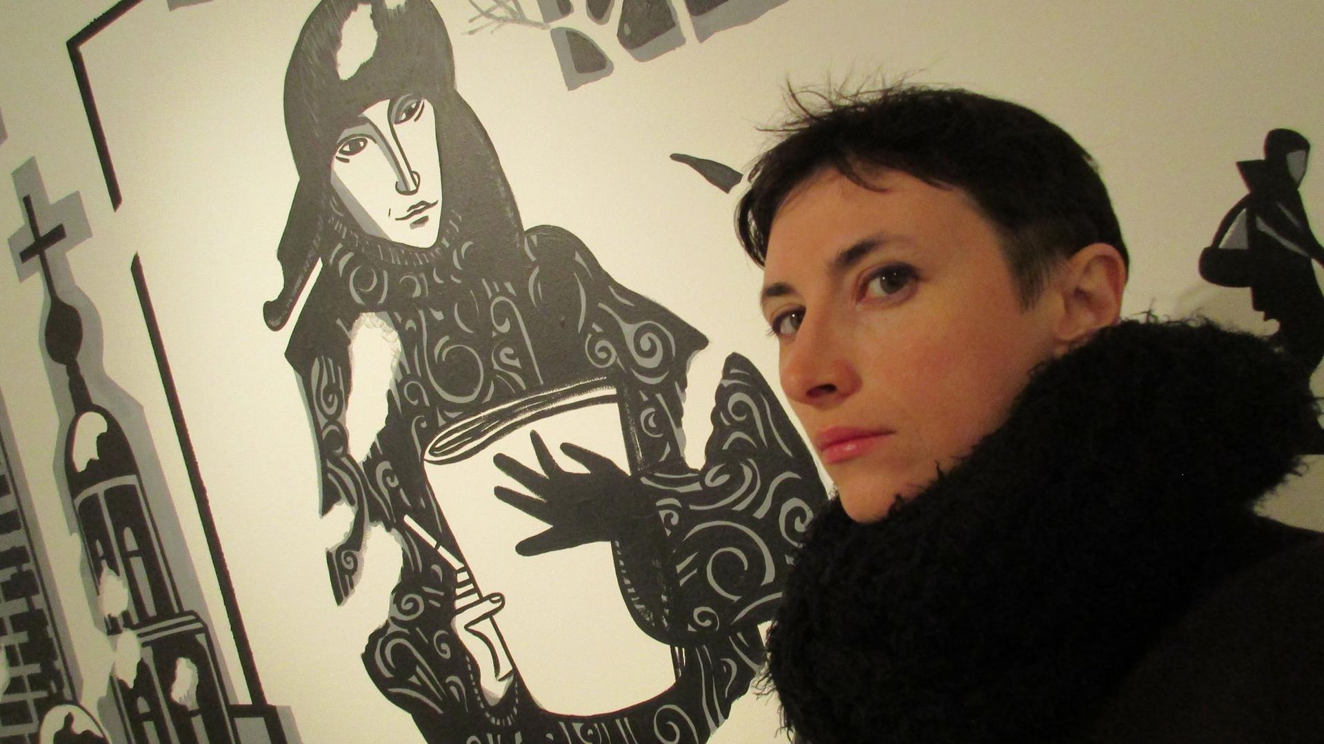 Victoria Lomasko is shown in this portrait photo with black coat standing in front of an illustrated wall.