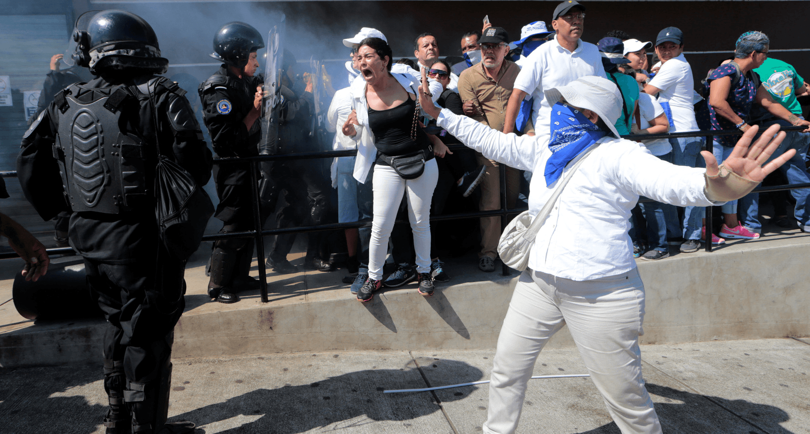 A woman wearing a blue bandana covering her face dressed in white holds her hands out to block a riot police. 