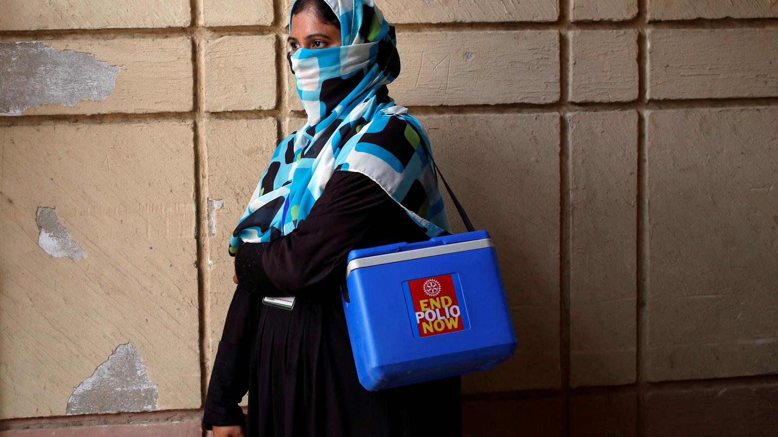 A woman wearing a scarf over her face stands by a wall carrying a polio vaccination kit box in Pakistan