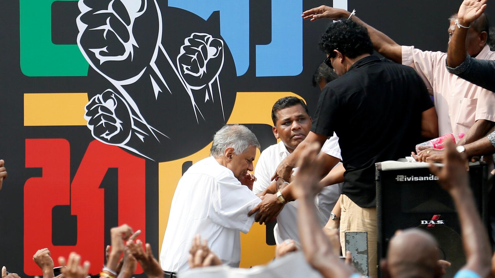 Prime Minister Ranil Wickremesinghe steps onto stage in front of colorful mural with black fist symbols 