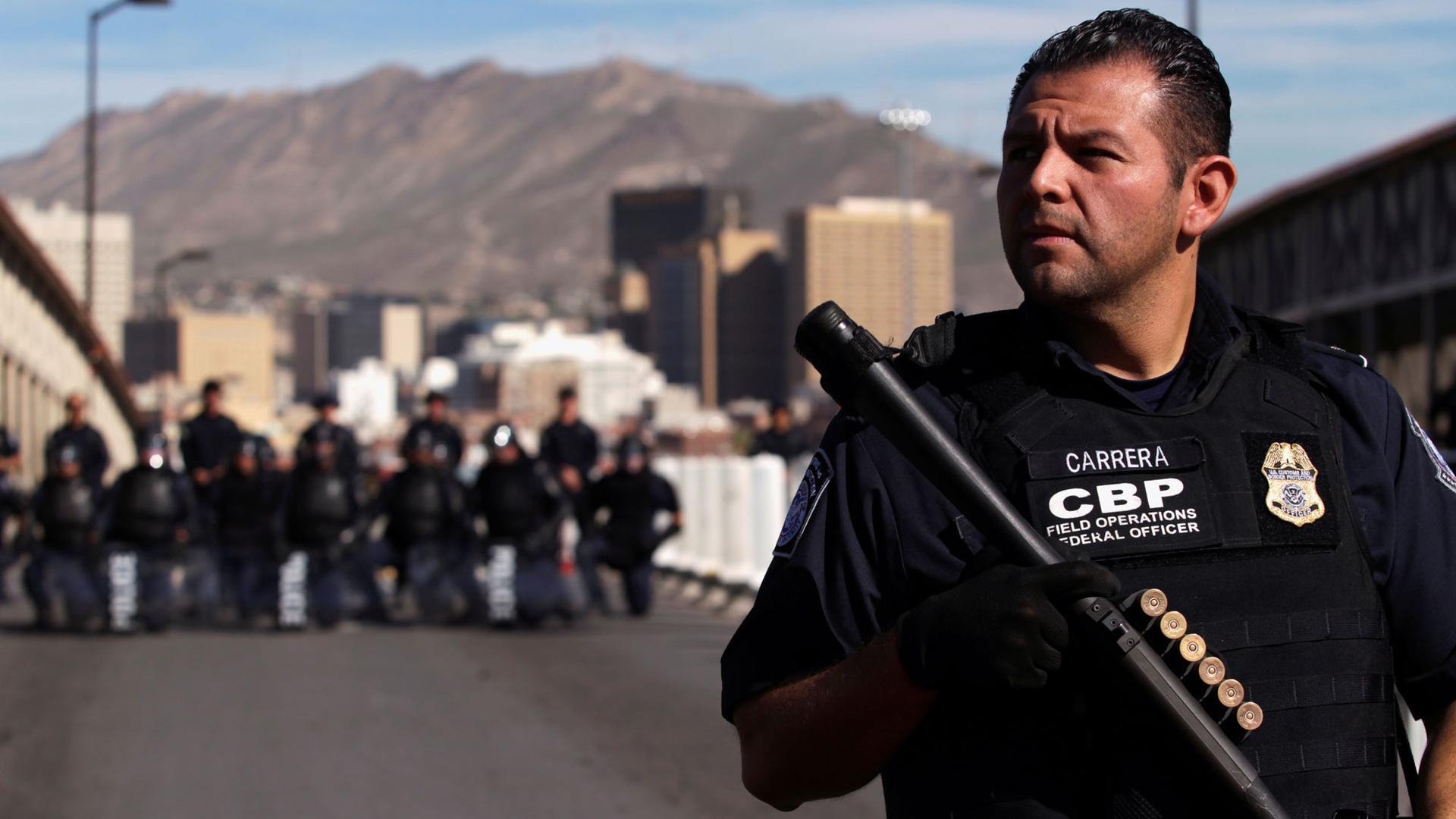 A US Custom and Border Protection agent stands in the foreground with a riffle additional agents armed in the distance.