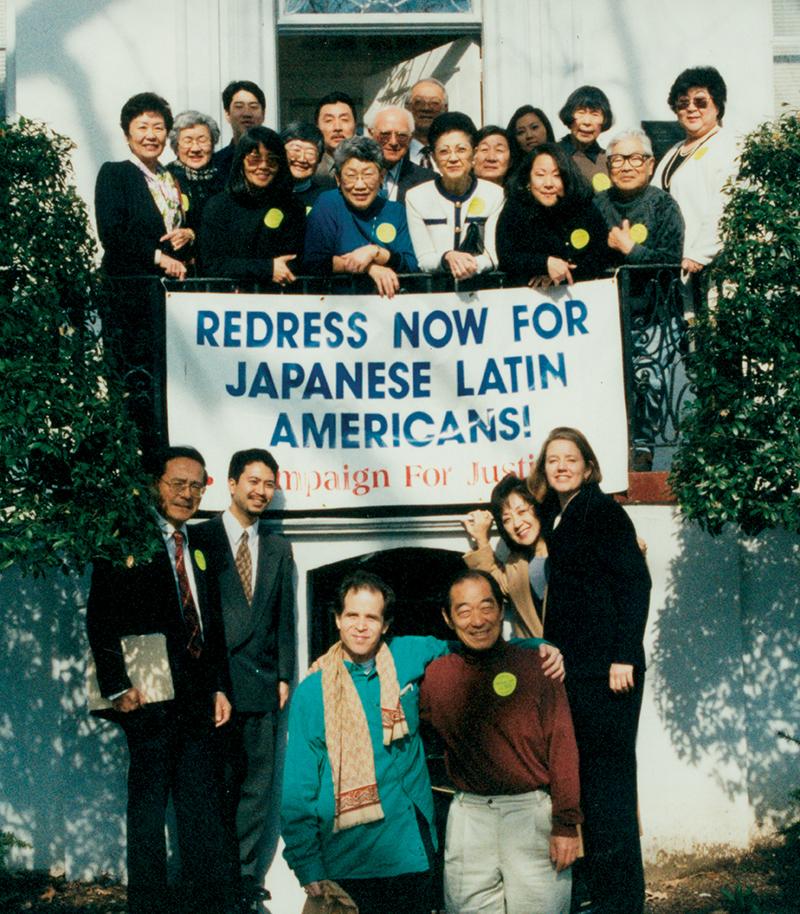 A group of men and women stand on a balcony above a banner reading 