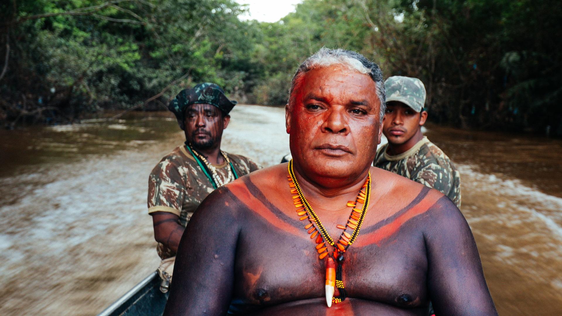 An Indigenous Man wearing beads, his chest decorated with red and black paint, sits in a speedboat. There are two men in camouflage behind behind him.