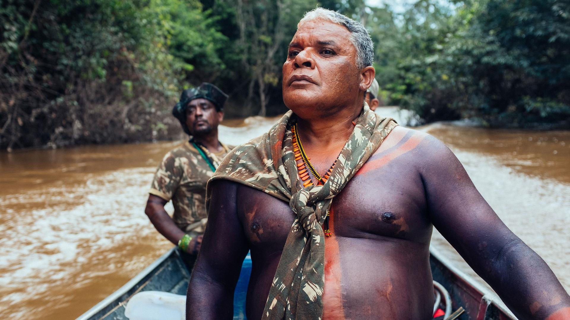 A barechested man wearing beads and body paint sits in a motorboat as it speeds through a muddy river