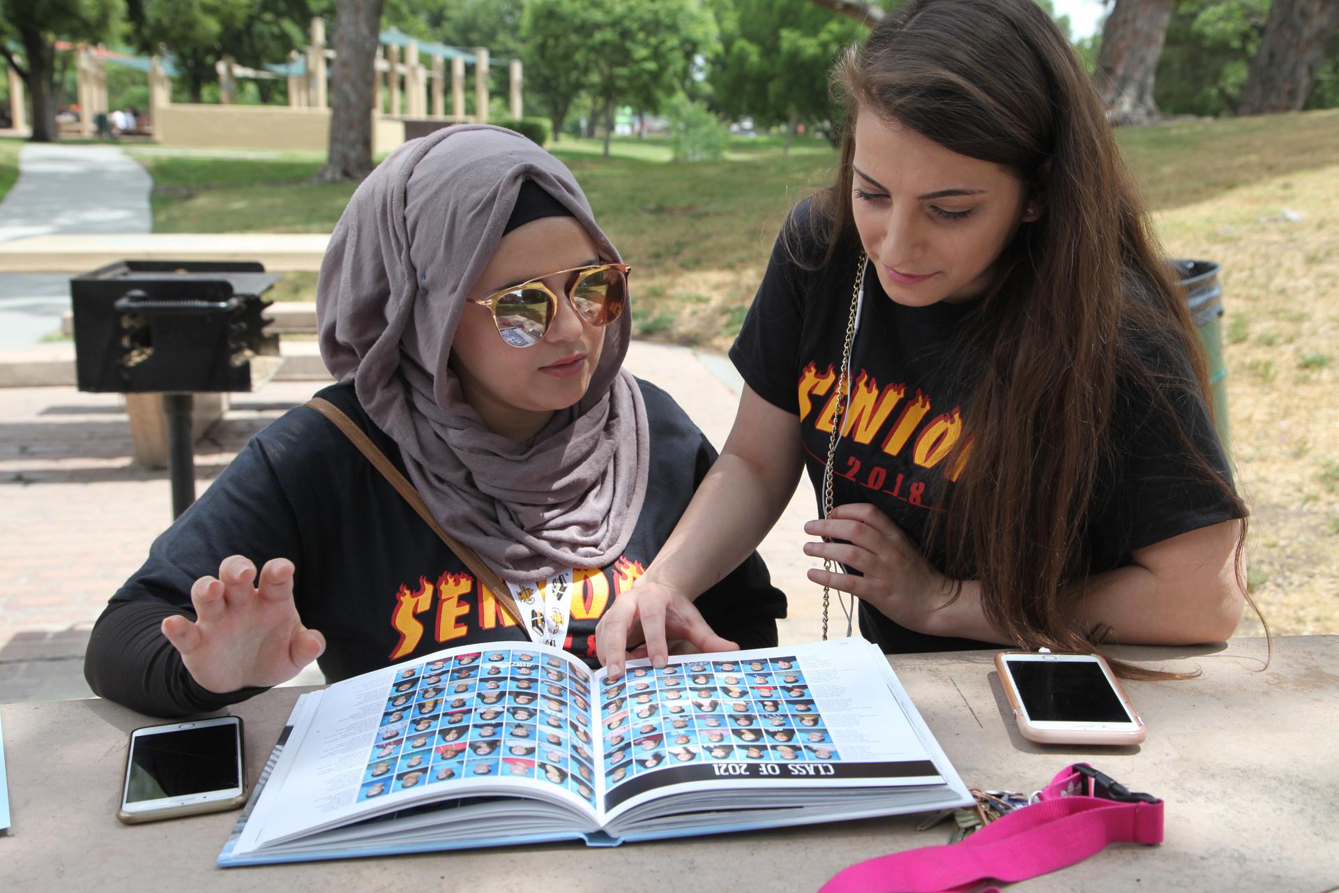Young woman sits at bench, flipping through year book, while another young woman stands next to her, leaning over to look