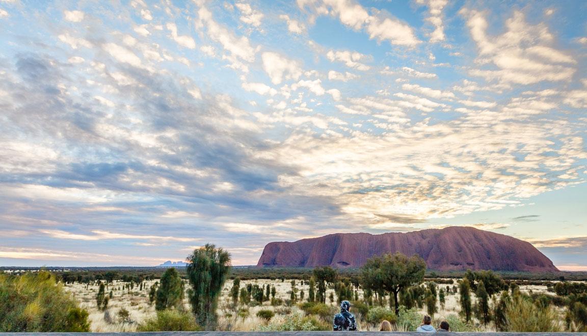 The Uluru rock formation is seen off in the distance with the sun rising across the sky.