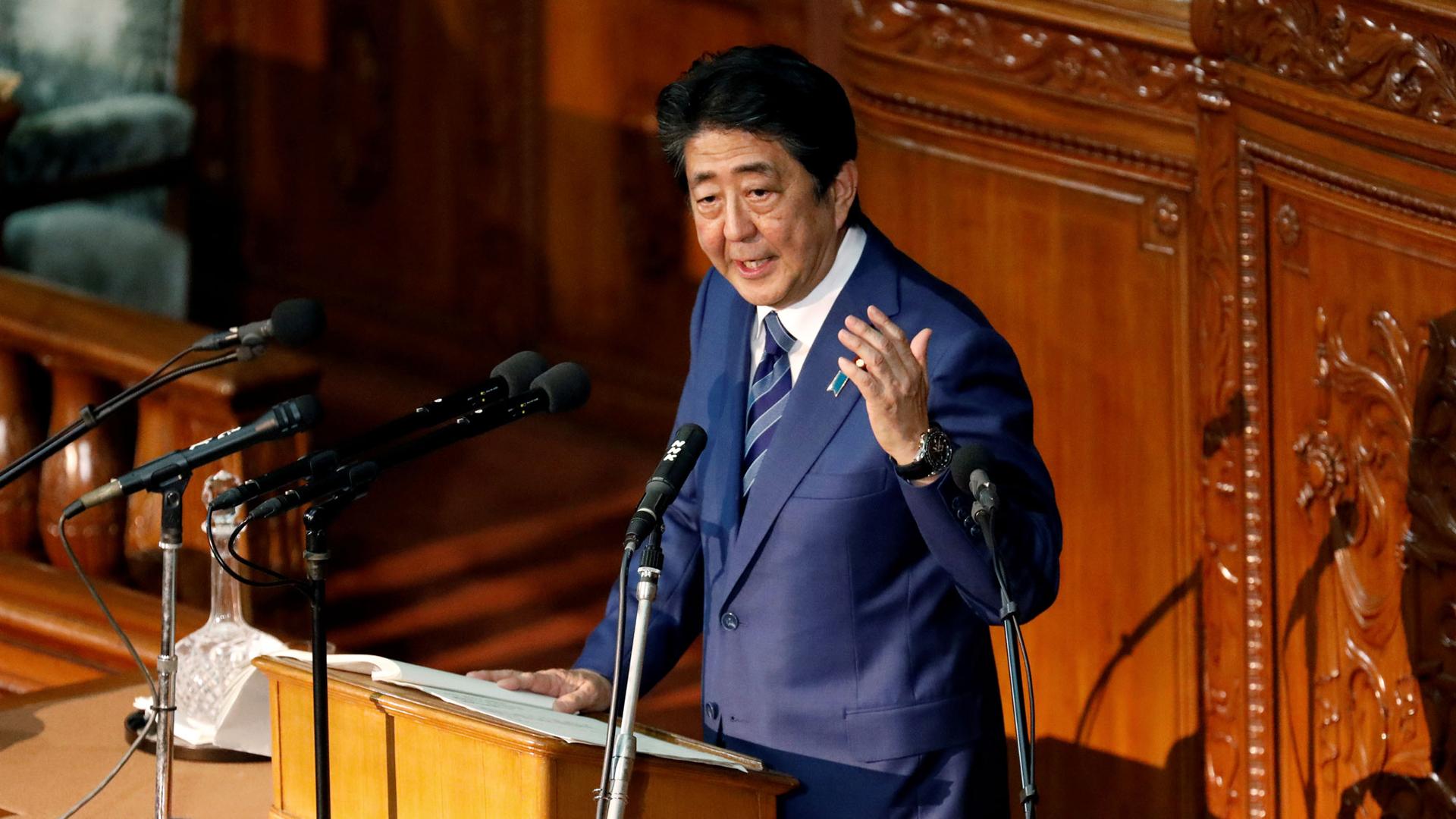 Japan's Prime Minister Shinzo Abe is shown standing at a podium with his left hand raised delivering a policy speech.
