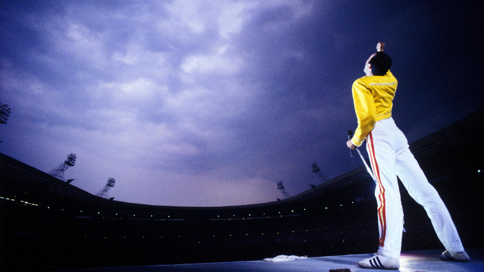 Freddie Mercury on stage wearing a yellow leather jacket and singing to the sky.