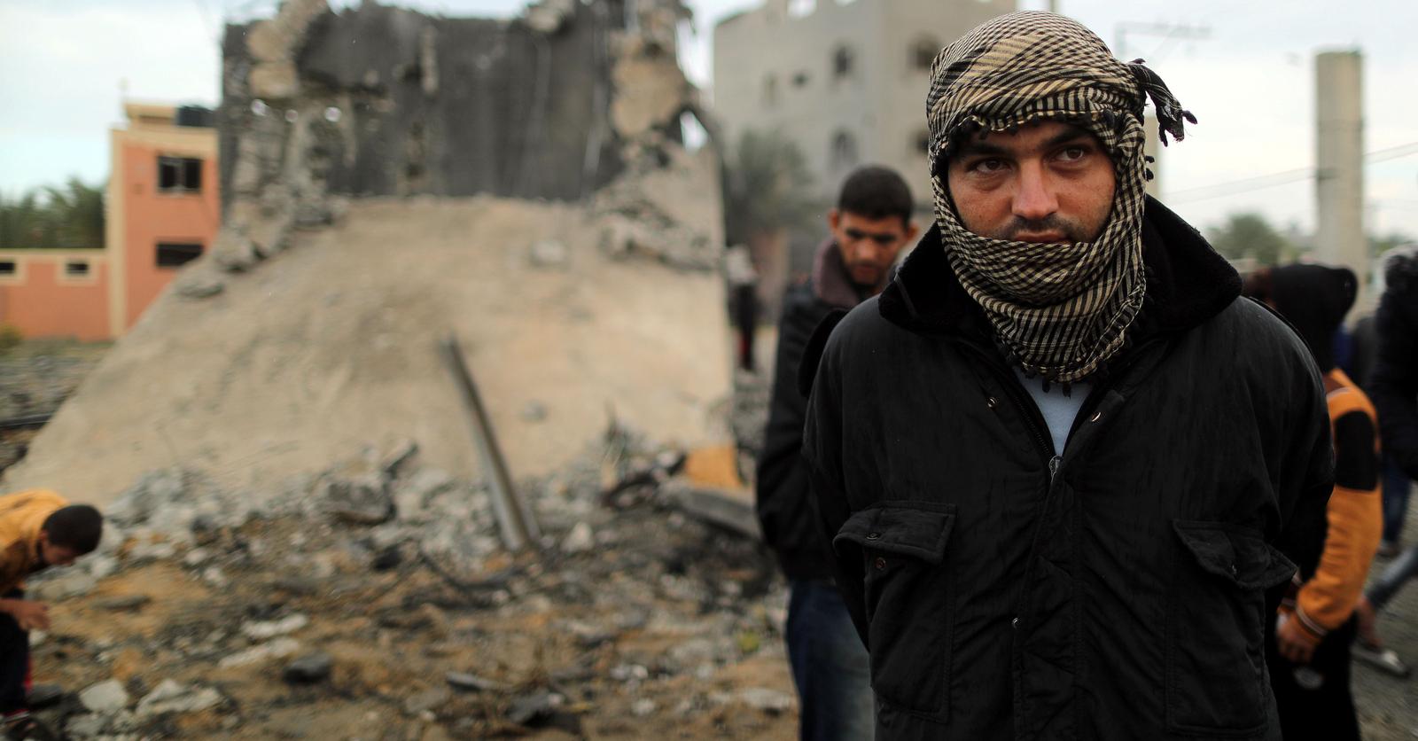 A Palestinian man wears a scarf and looks on as he stands in front of a building that was destroyed by an Israeli air strike. 