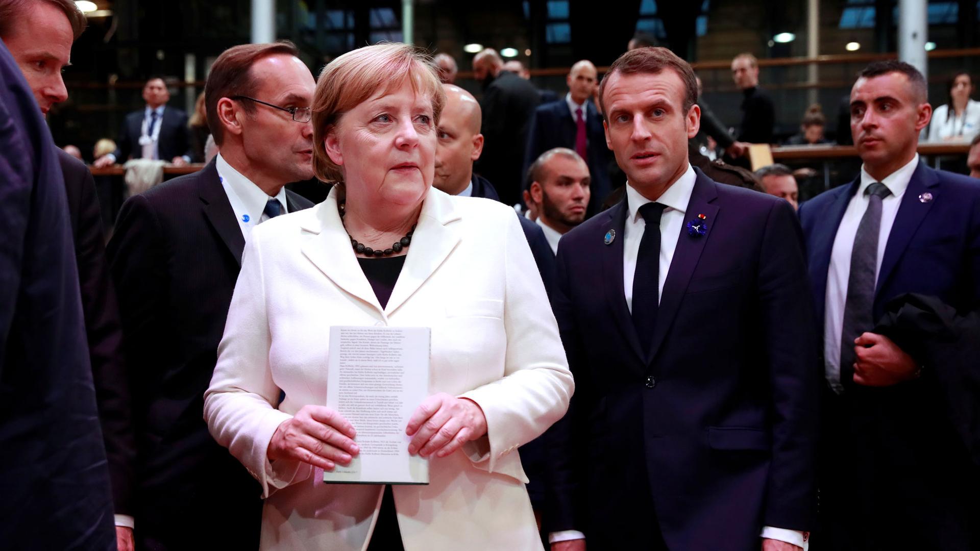German Chancellor Angela Merkel, wearing a white jacket, and French President Emmanuel Macron, in a dark suit, are shown standing next to each other at the Paris Peace Forum.