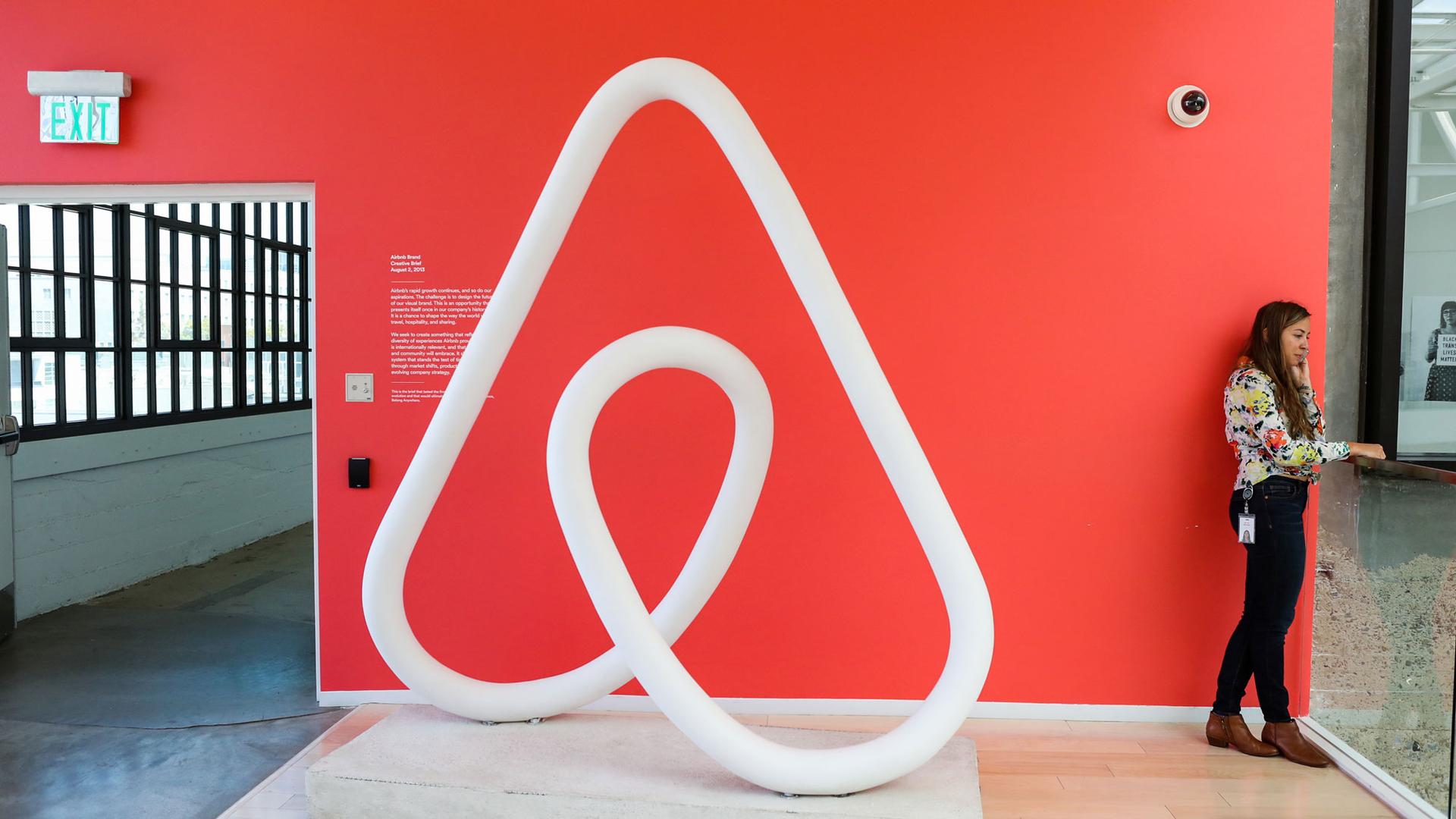 A woman is shown talking on the phone next to a red wall and a large sculpture of the Airbnb logo at the company's headquarters in San Francisco, California.