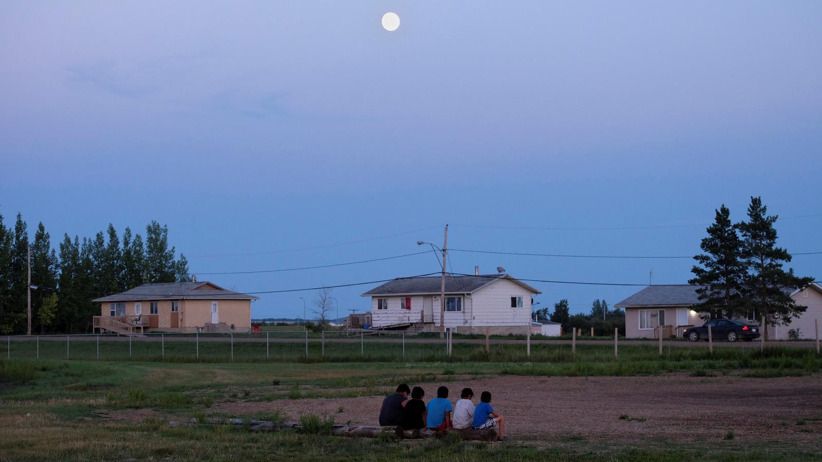 Children sit on a log under the moonlight in a field surrounded by buildings.