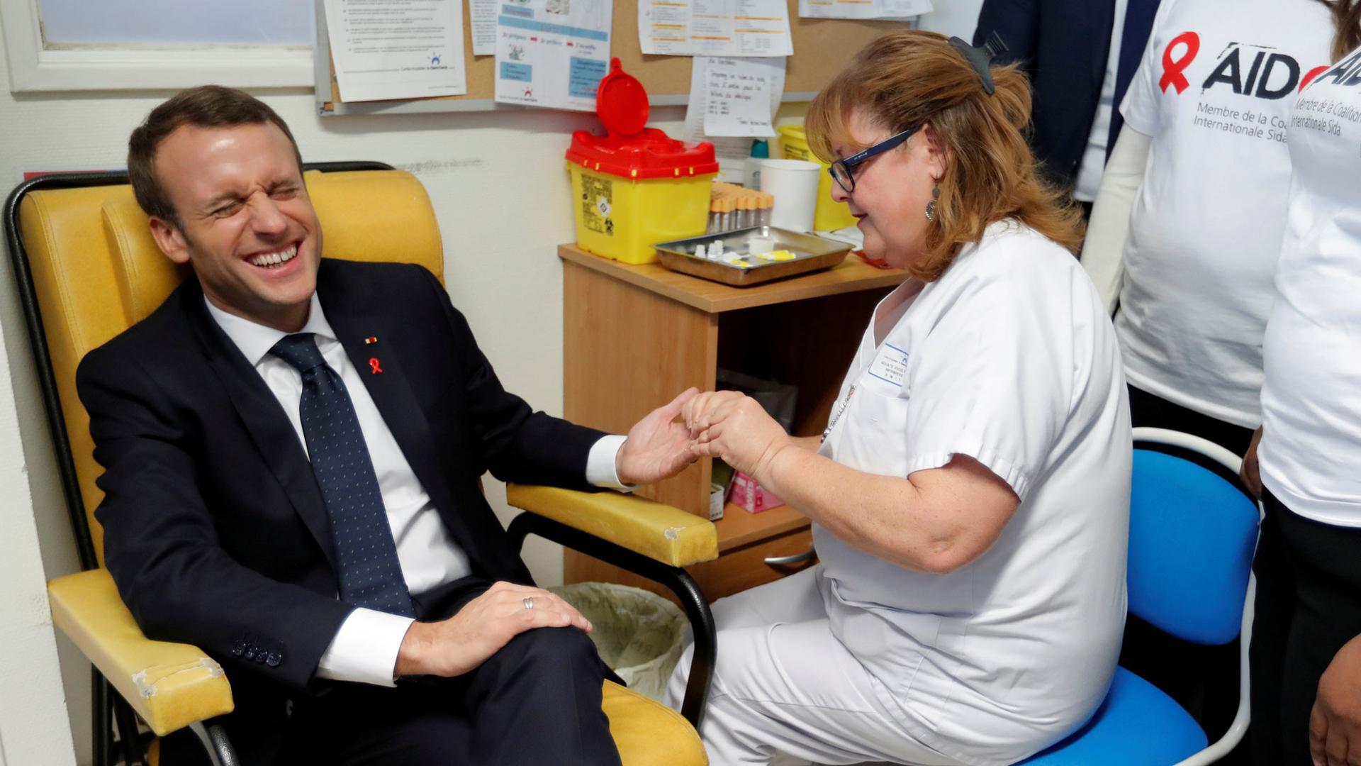 A nurse is shown taking blood for a HIV test from French President Emmanuel Macron who is sitting in a yellow chair cringing.