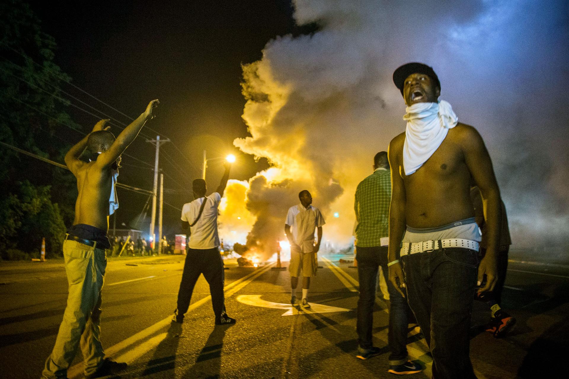 Men stand under the glow of a street light in the street with their hands up. In the distance is a cloud of tear gas.