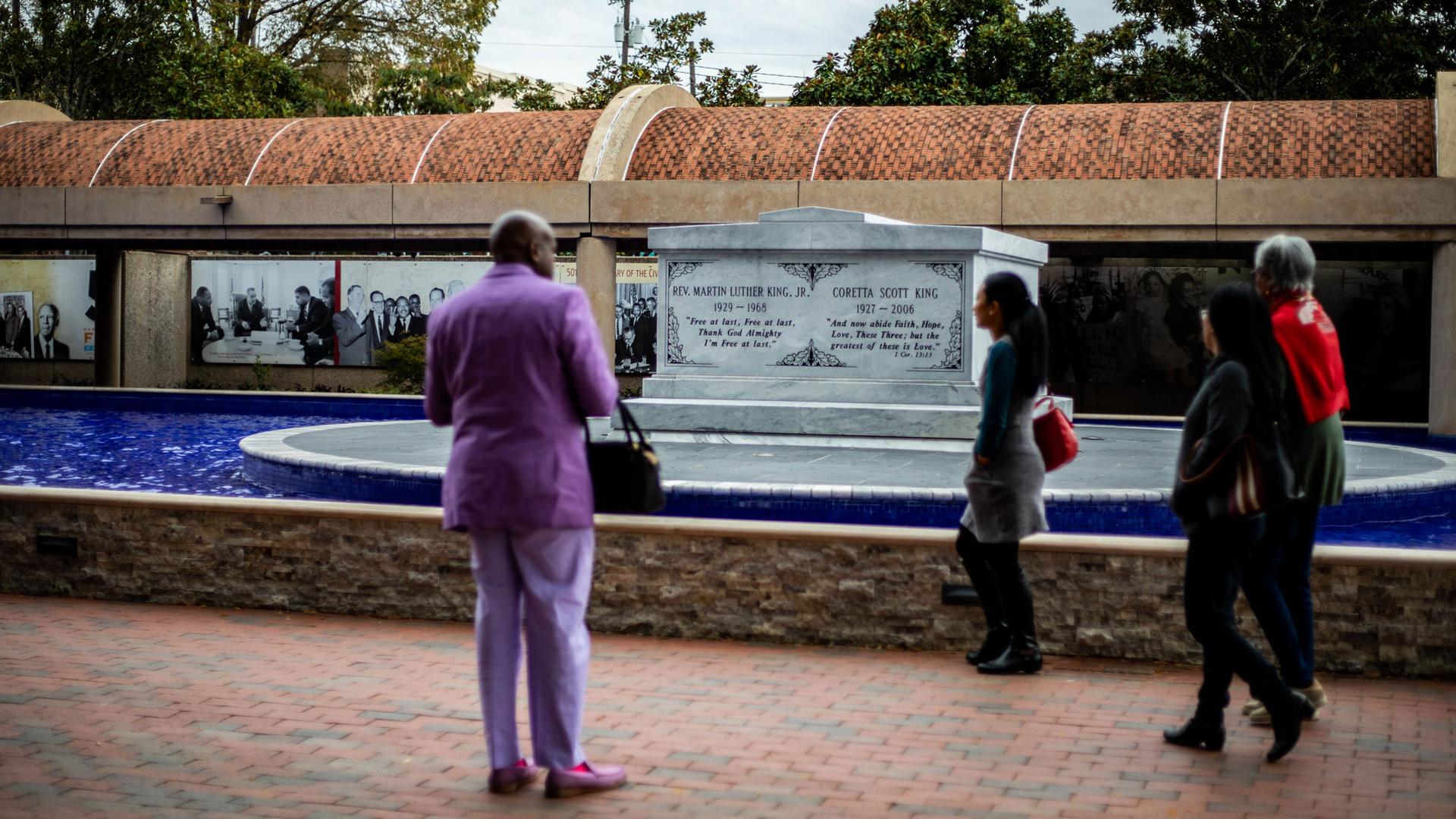 Several people including a man wearing a purple suit, look on at the stone monument for Martin Luther King, Jr. 