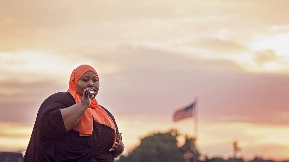 A woman in a hijab speaks into a microphone as dawn breaks in the clouds. An American flag waves in the distance.