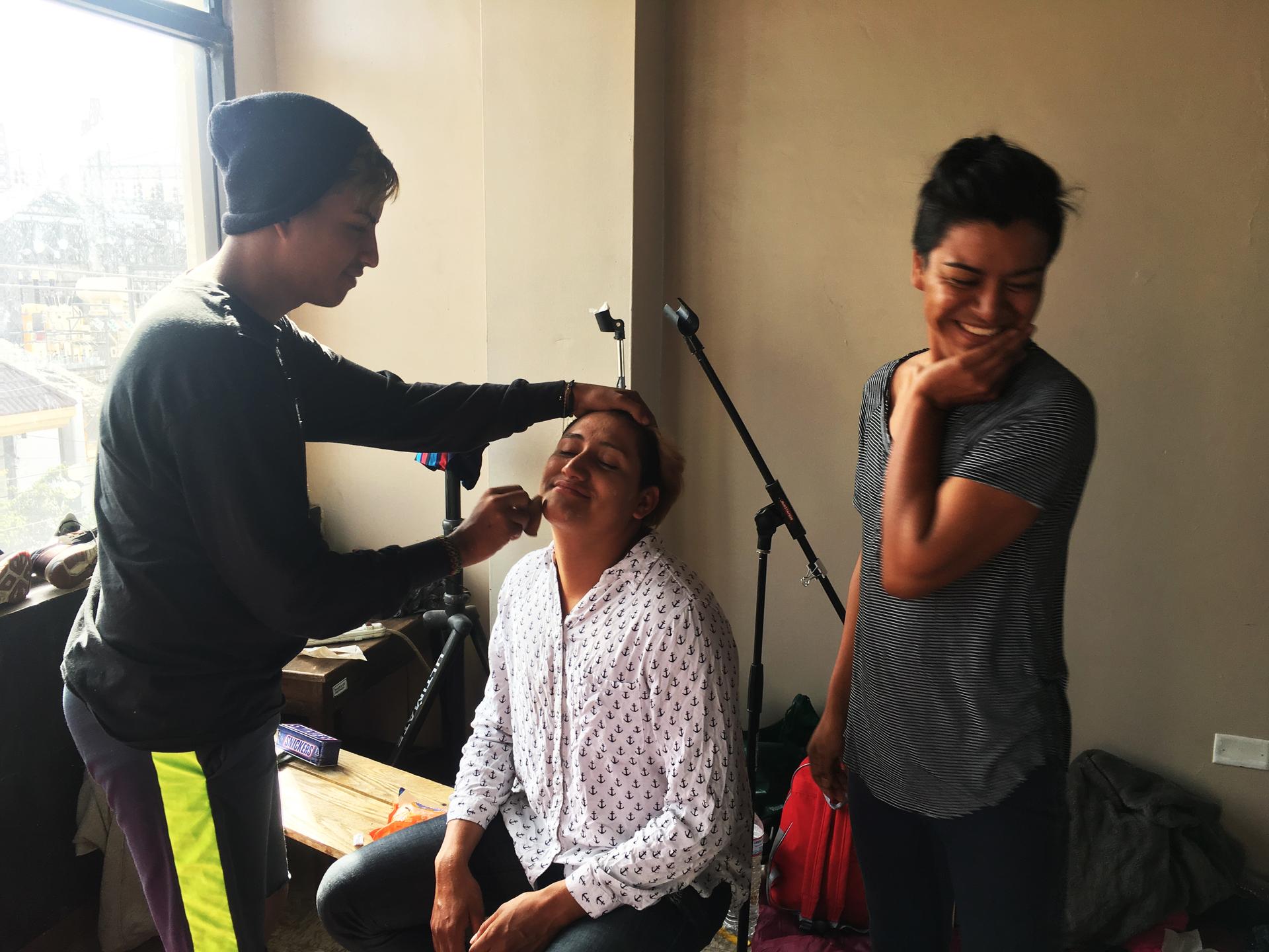 A few migrants prepare for the weddings by putting on makeup. One sits in a chair while another applies makeup and another giggles in the corner