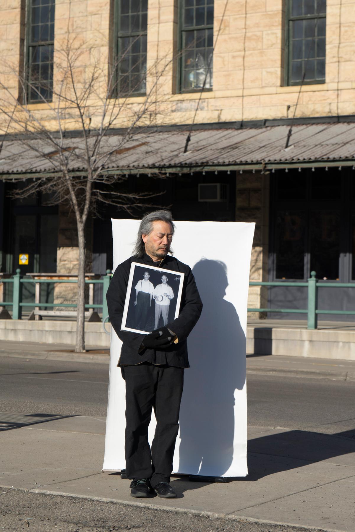 A man stands outside holding a photograph in front of a white sheet