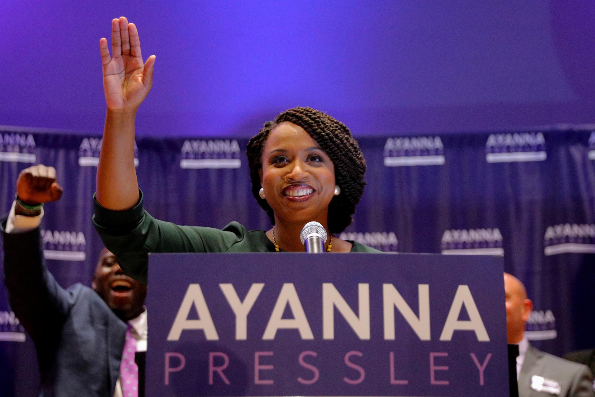 Representative-elect Ayanna Pressley stands behind a podium and waves to the crowd as her husband cheers behind her